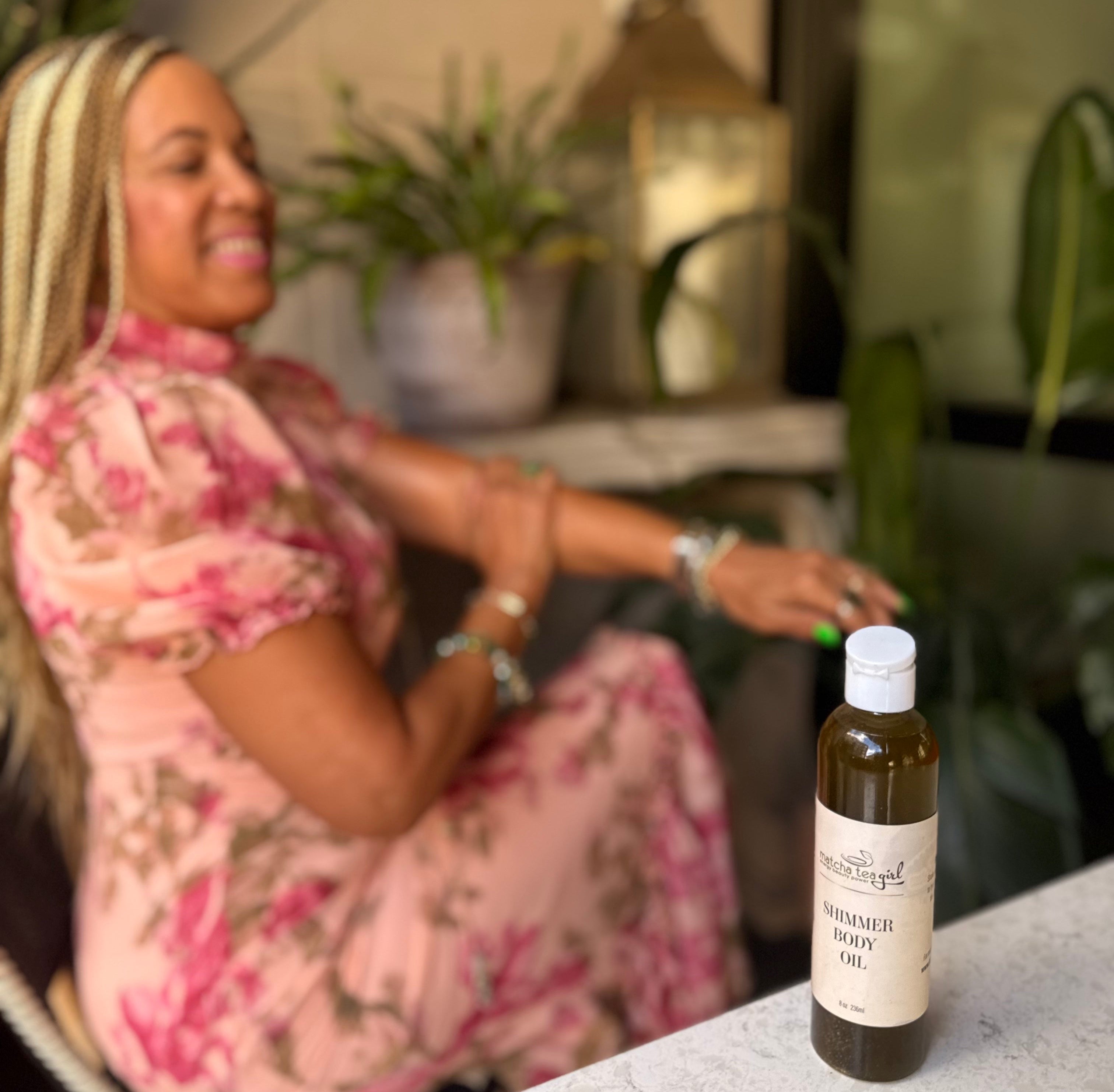 Woman in a floral dress sitting outdoors with a bottle of body oil on a table in front of her. She's rubbing the body oil on to her skin. 